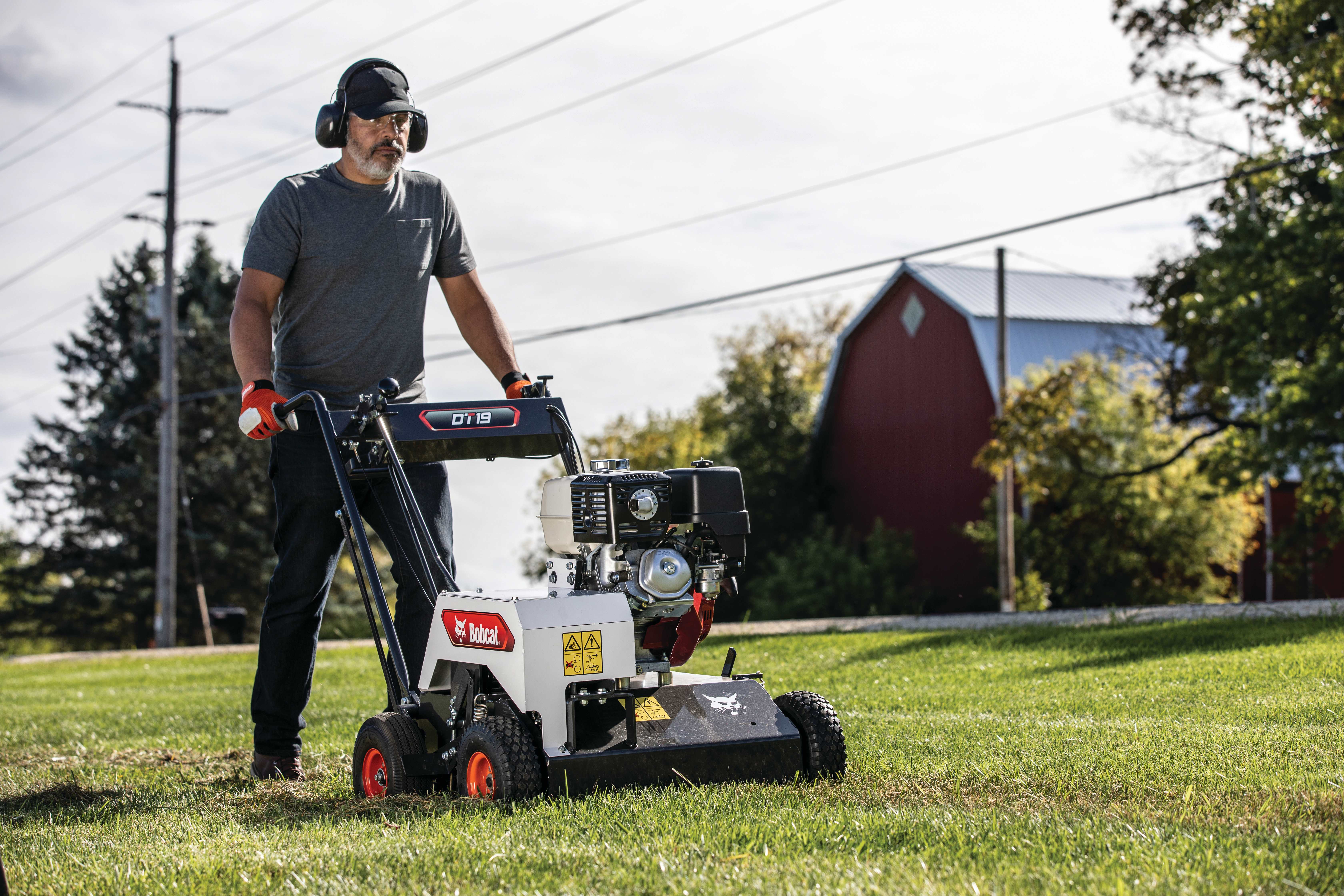 Male operator dethatching lawn with Bobcat DT19 dethatcher with large red barn and trees in the background