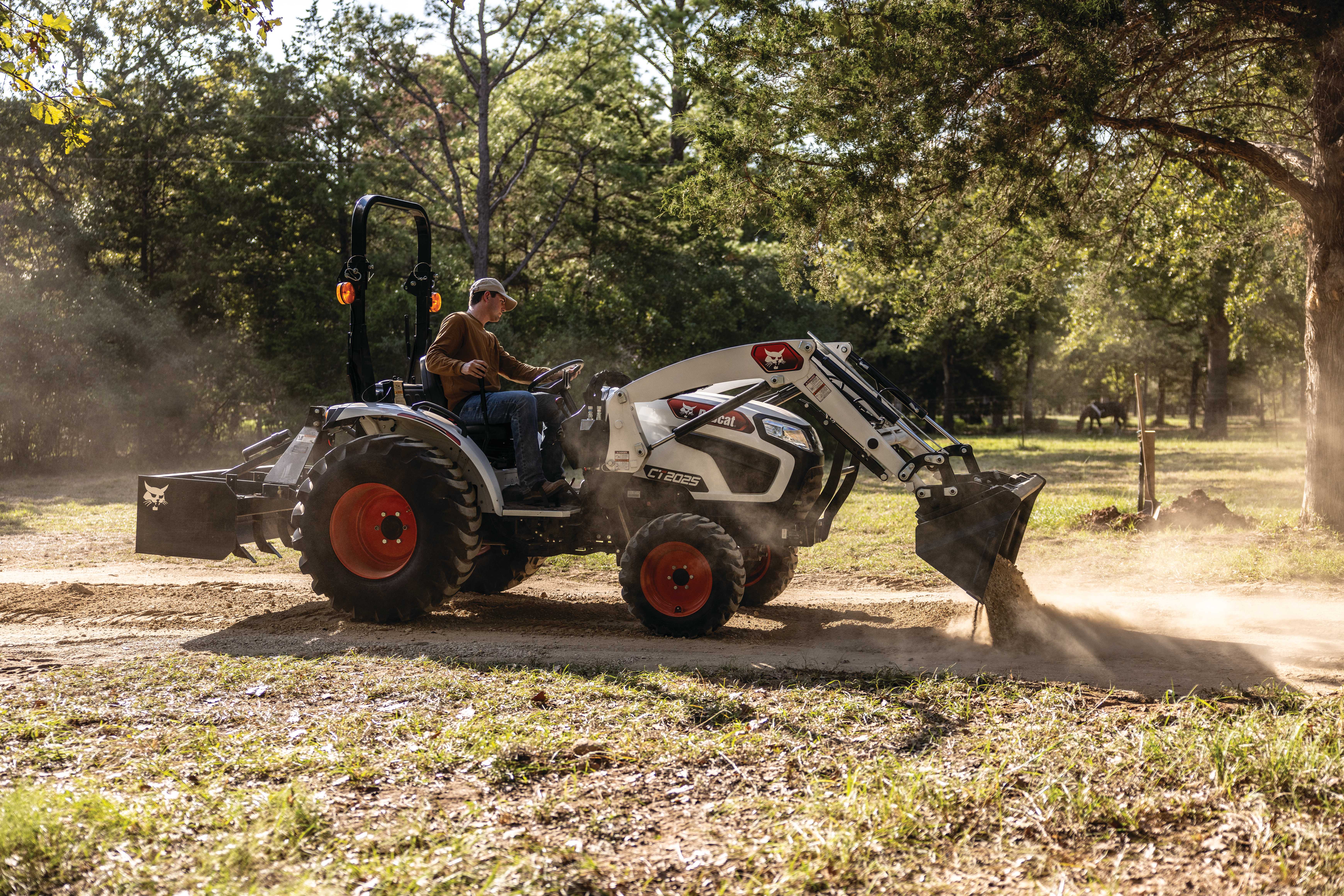 Bobcat CT2025 compact tractor with bucket attachment dumping a load of sand on a trail with trees and pasture in the background