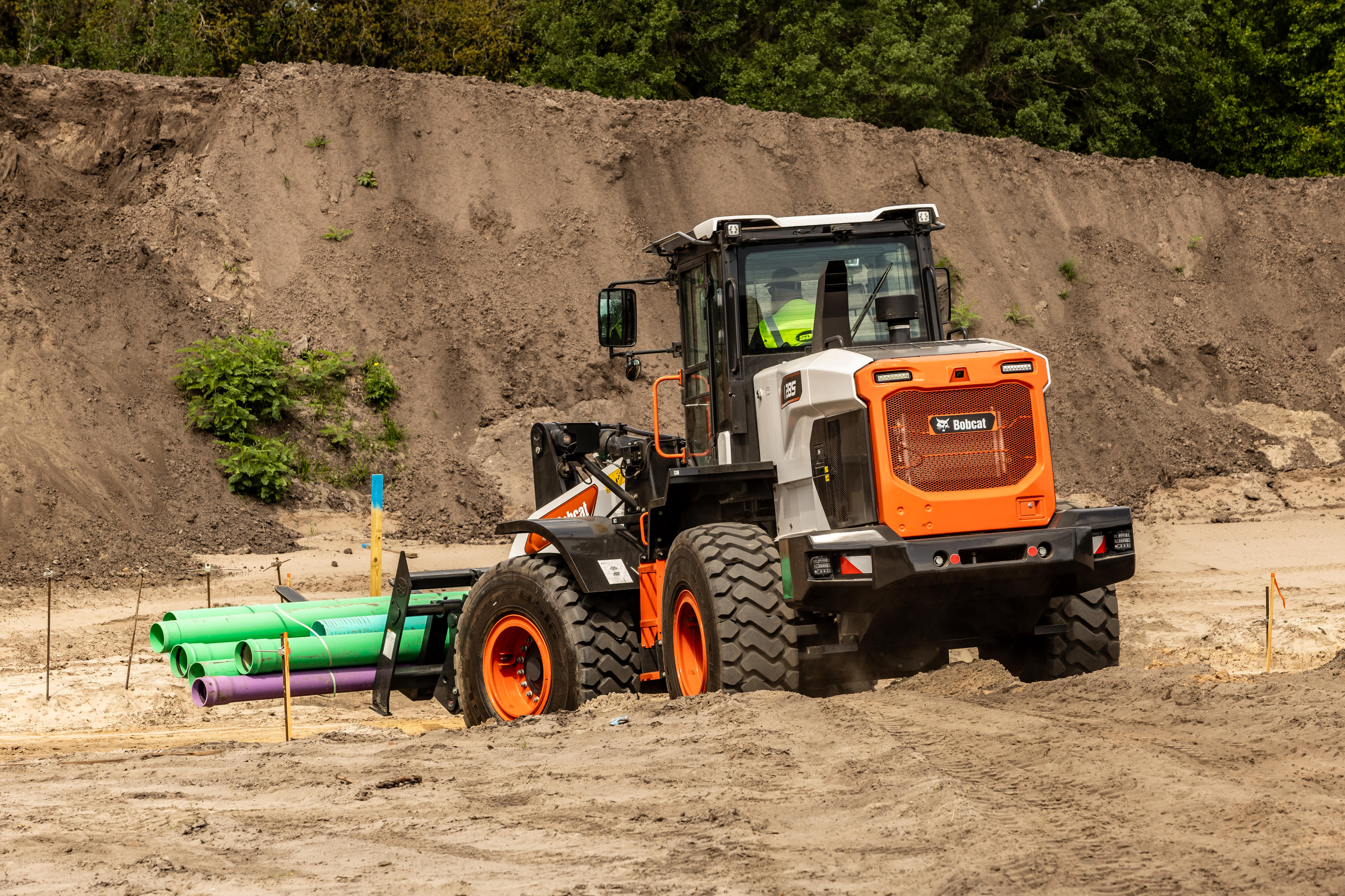 Bobcat L285 Large Wheel Loader with Pallet Fork Attachment Carrying Utility Pipes Across a Sandy Construction Site