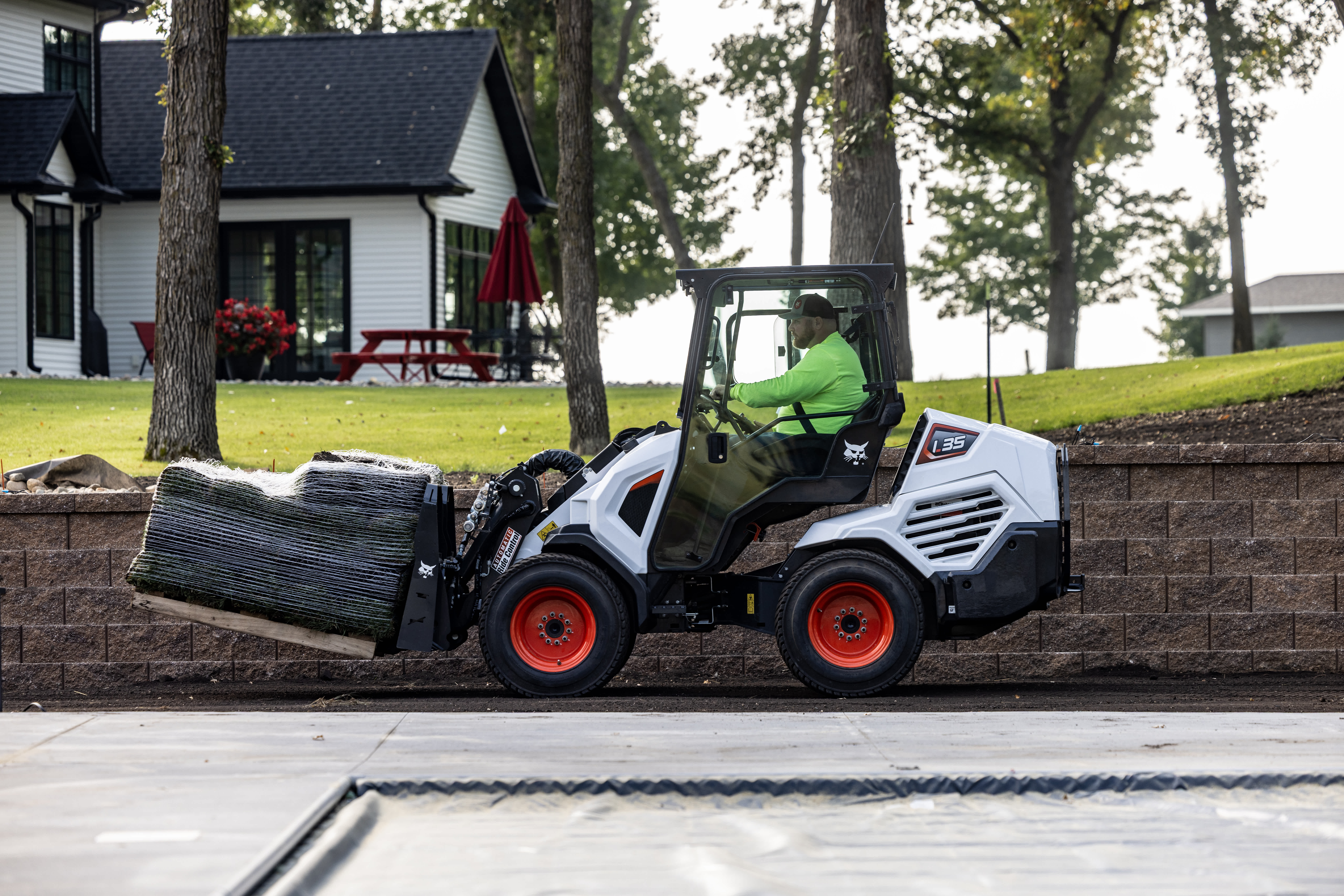 Side profile view of Bobcat L35 small articulated loader traveling on jobsite carrying a pallet of sod rolls with white home and yard with trees in the background
