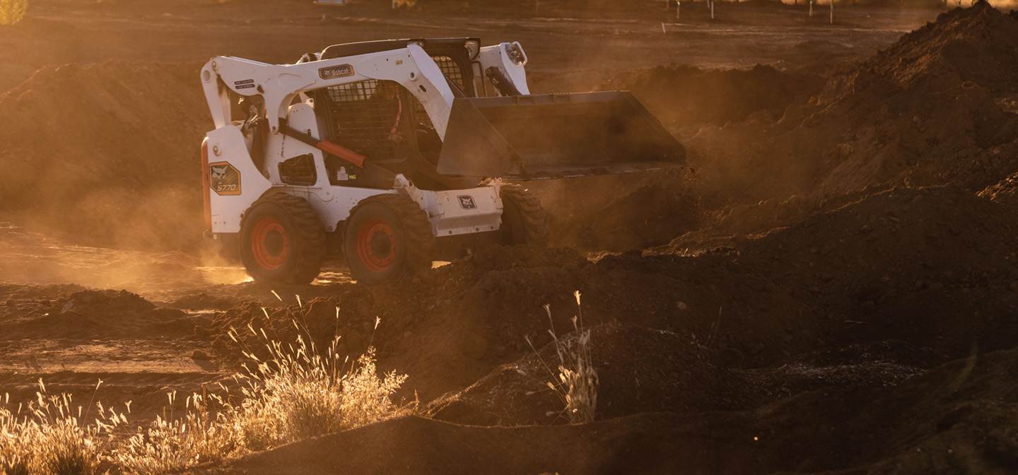 A Bobcat Classic S770 Skid-Steer Loader Uses a Bucket to Move Dirt on a Jobsite During Sunset With Dust in the Air
