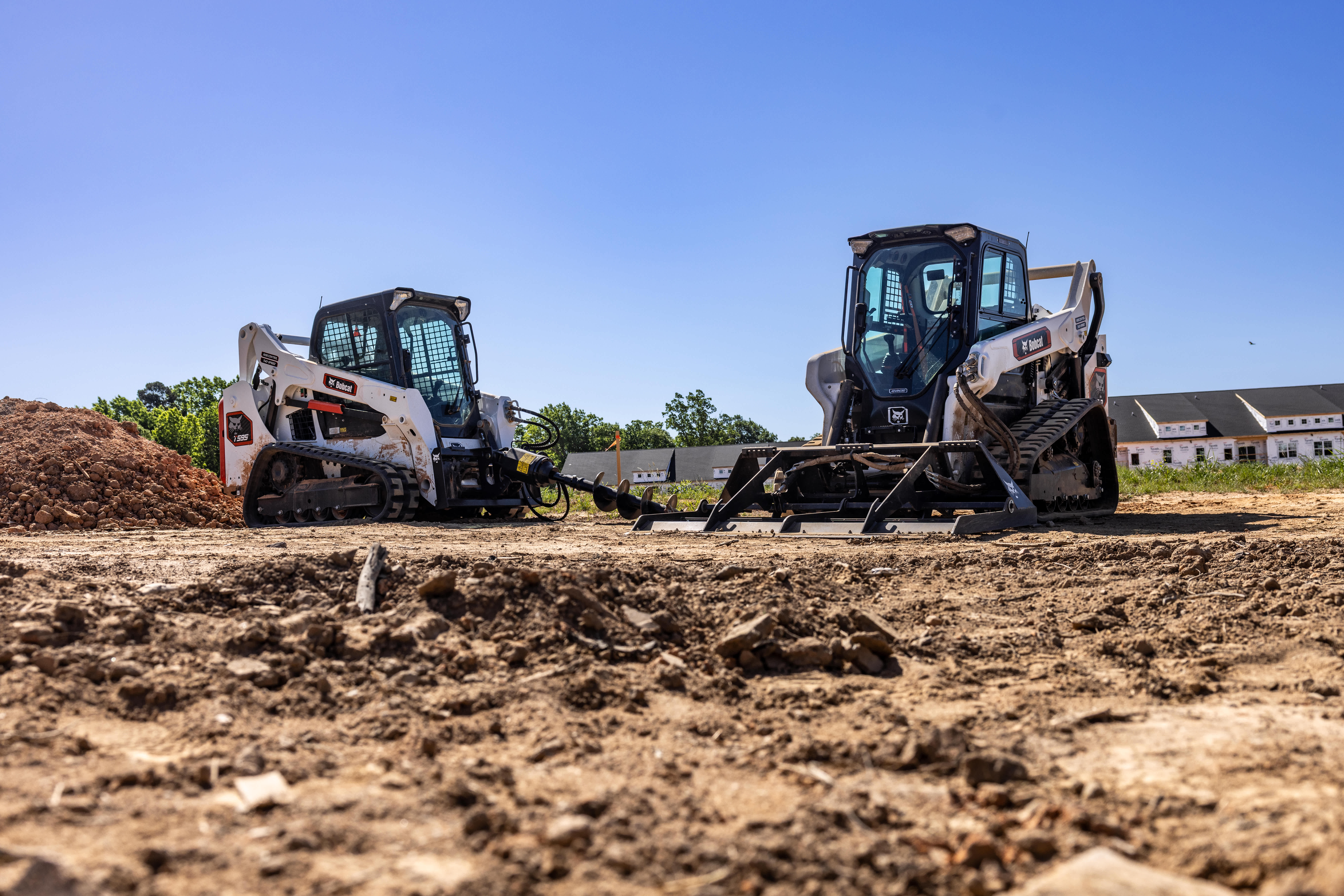 Bobcat T595 Compact Track Loader With an Auger Attachment Positioned Beside a Bobcat T66  Compact Track Loader With a Landplane Attachment at a Residential Construction Site