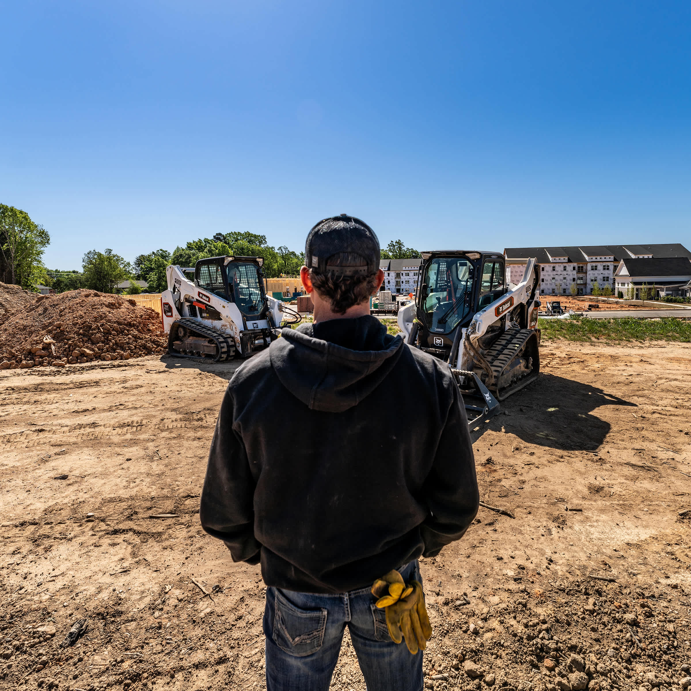 An Operator Stands Near a Bobcat Pro T66-2 and Classic T595 Compact Track Loader at a Residential Construction Site With Dirt Piles and New Homes in the Background