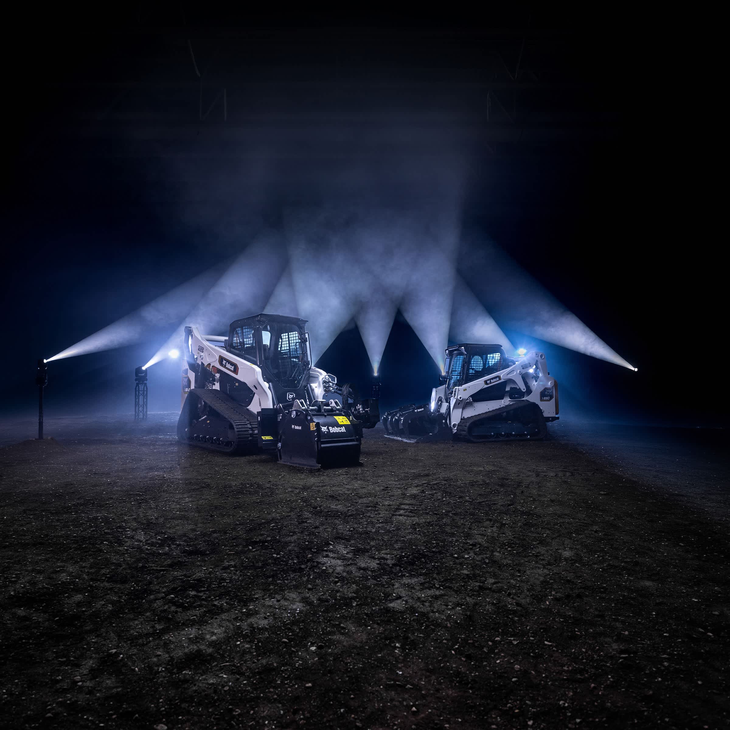 A Bobcat T86-2 Compact Track Loader With a Planer Attachment and a Bobcat T650 Compact Track Loader With a Grapple Attachment Positioned at a Night Job Site With Dramatic Backlighting
