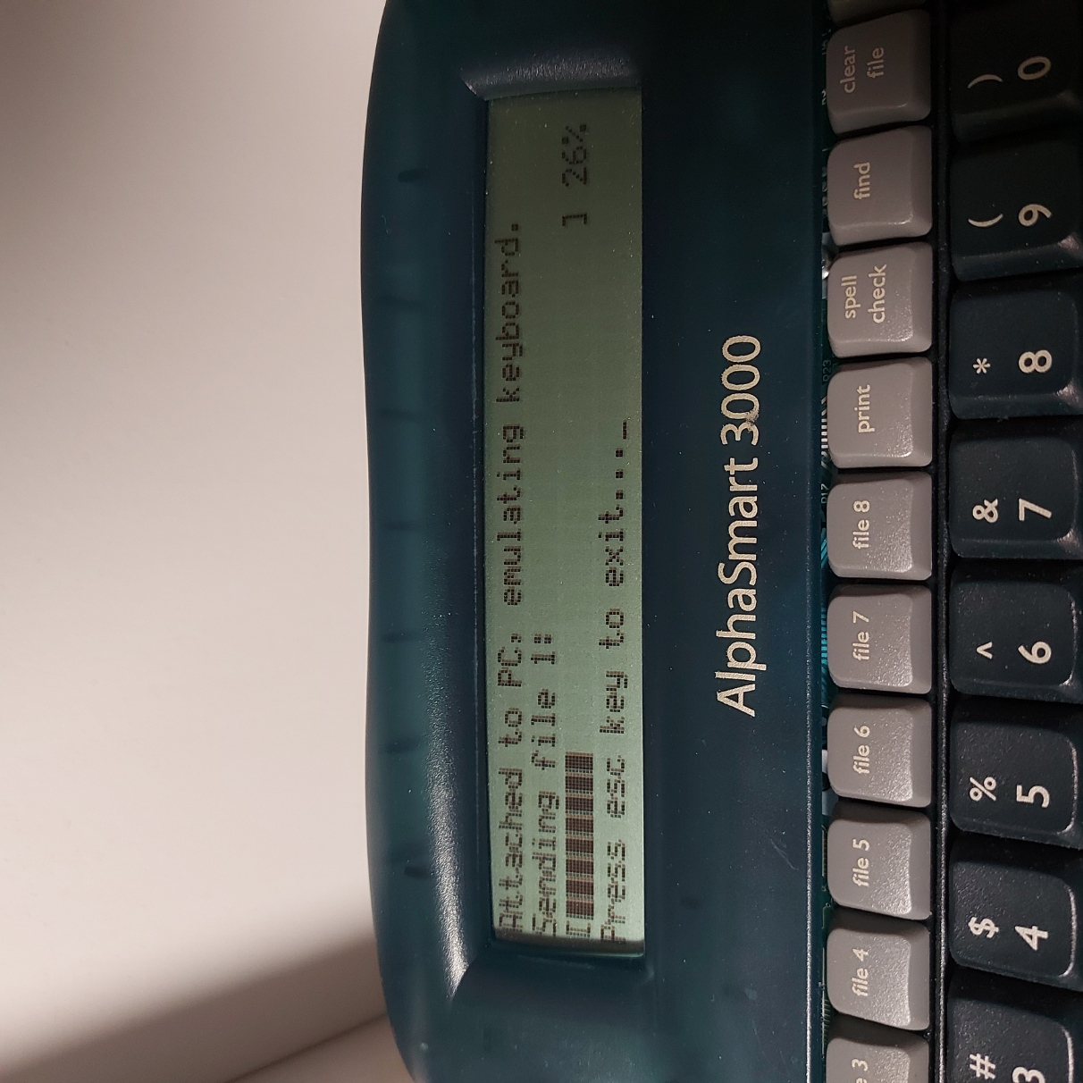 The screen of the AlphaSmart says 'Attached to PC, emulating keyboard. Sending file 1', followed by a progress bar showing 26%.