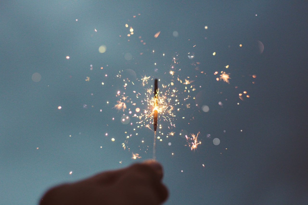 A hand holding a lit sparkler emitting sparkling sparks against a blurred blue background.