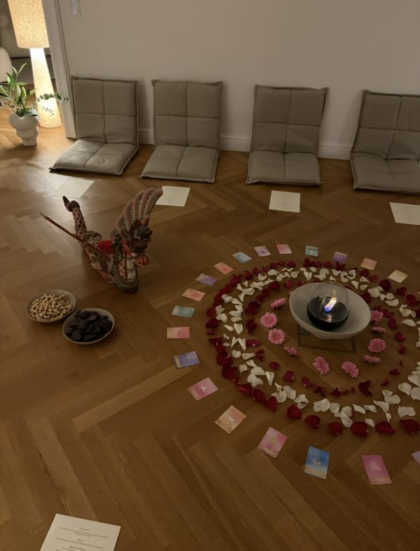 A circle of colorful cards, rose petals, and pink flowers surrounds a burning candle on a wooden floor, with four comfortable floor cushions and a decorative dragon figure nearby.