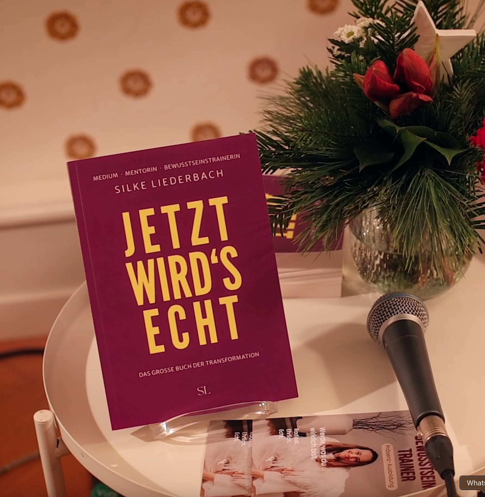 The book "Jetzt wird's echt" by Silke Liederbach is placed on a round white table next to a microphone and floral decoration.