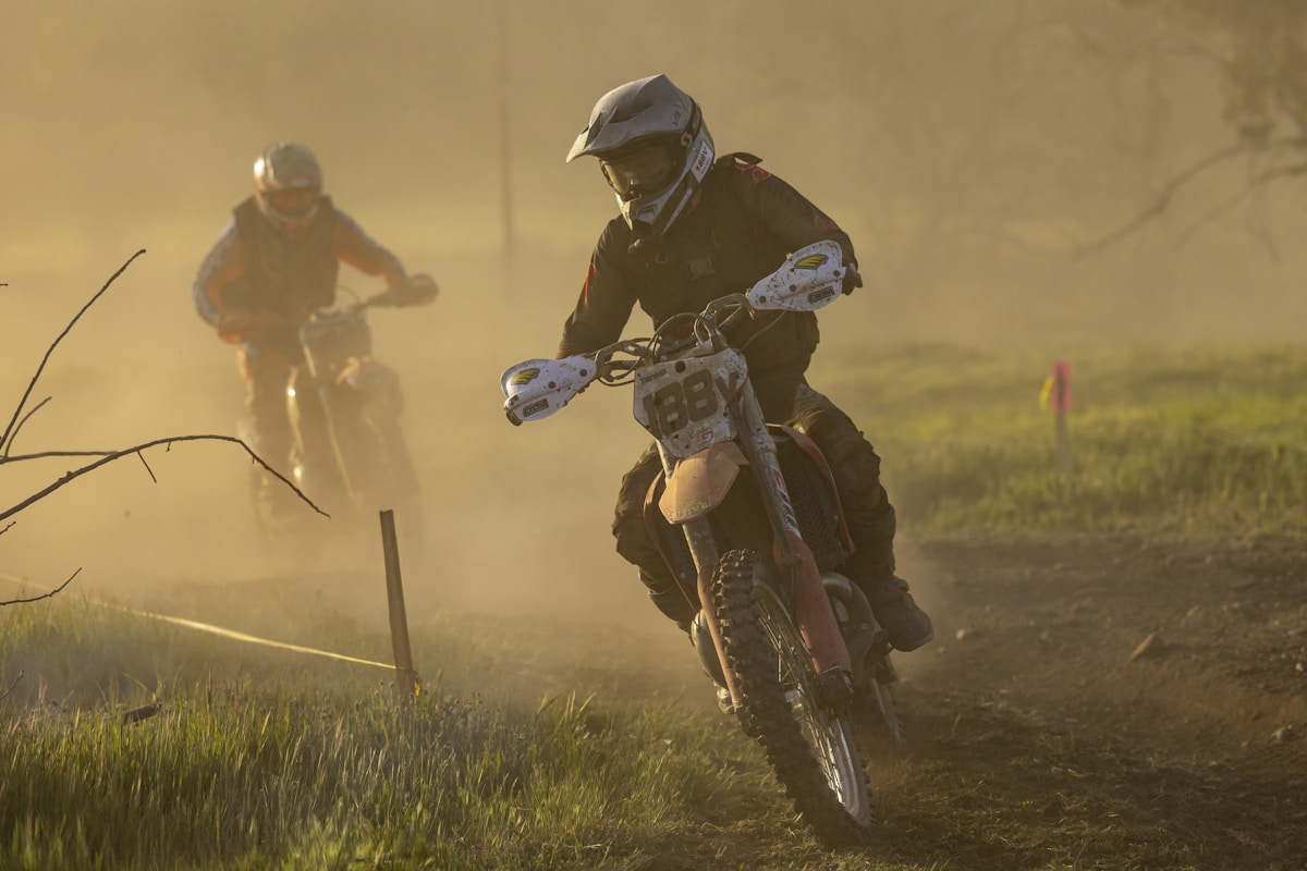 Motocross rider mid-air on dirt track at dusk, dramatic red dust clouds, dark rugged terrain with dynamic action lighting