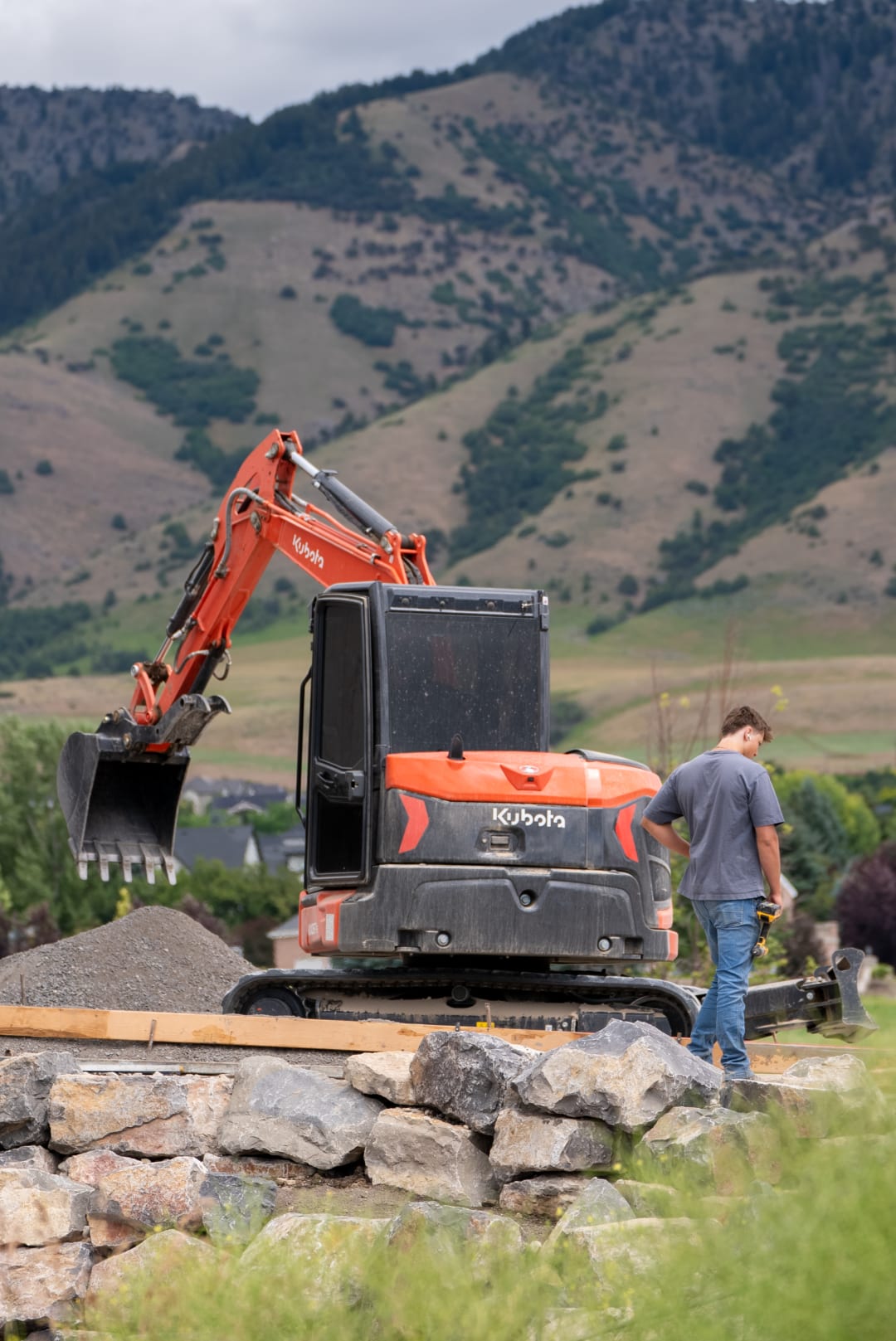 Kubota mini excavator setting boulders on a rock retaining wall course, foothills behind
