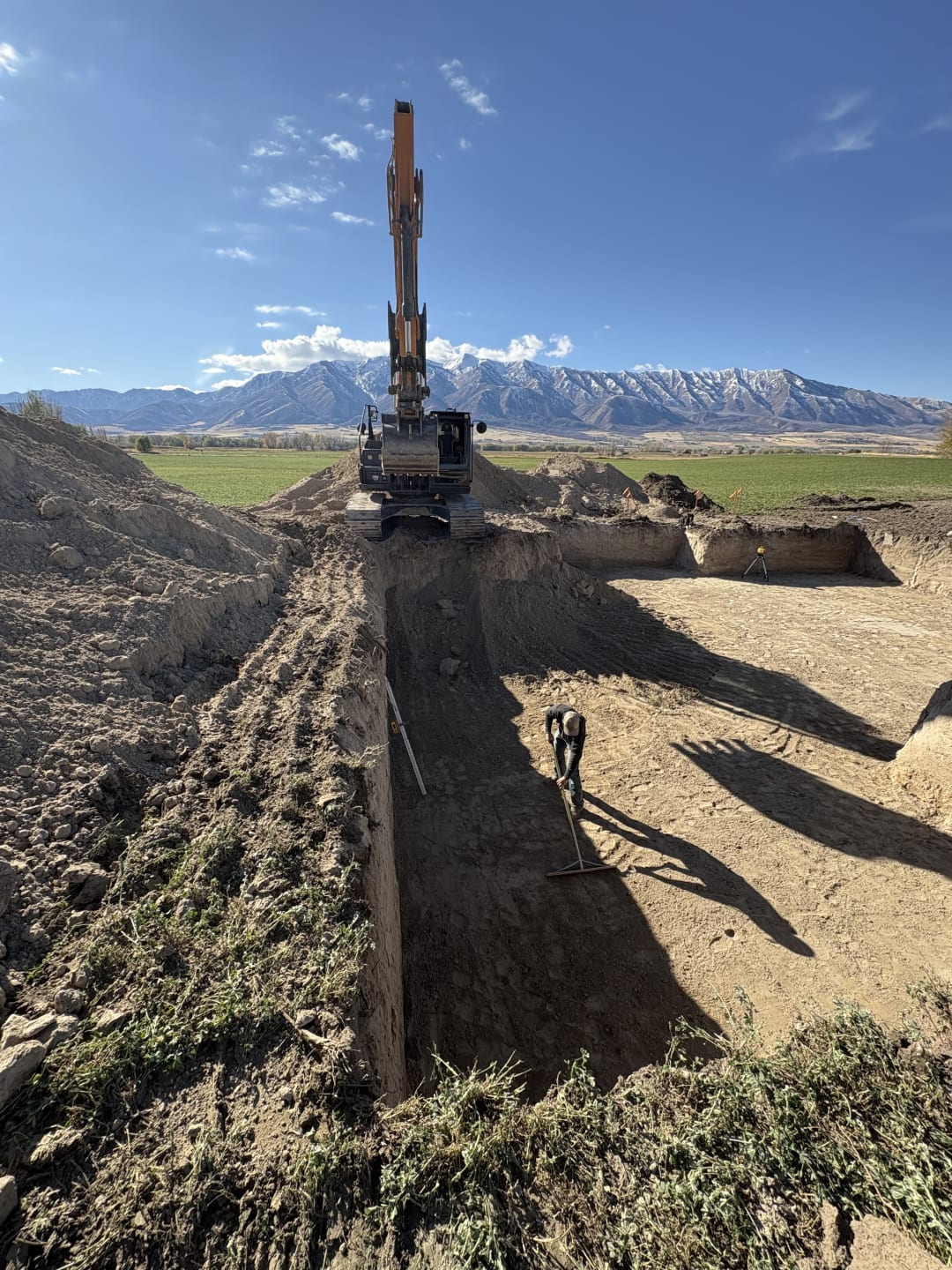 Excavator standing over a deep foundation cut with a worker raking the base, snow-covered mountains behind