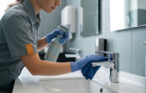 Person mit Handschuhen reinigt ein glänzendes Waschbecken mit Armatur in einem gewerblichen Sanitärraum