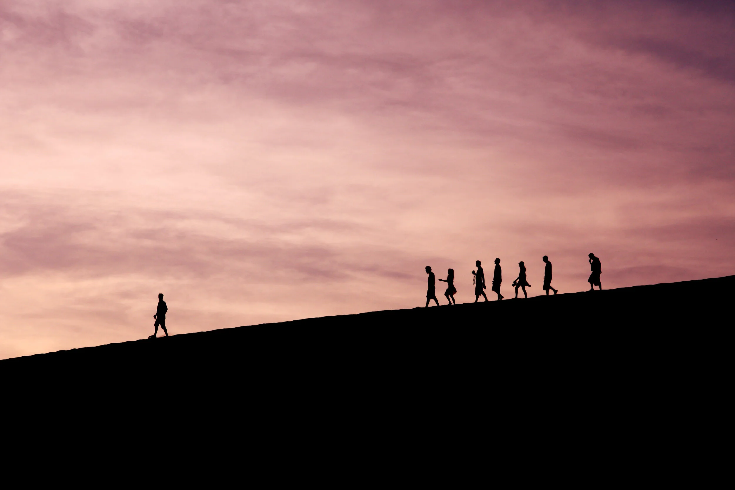 A silhouette of a person standing on a dune with other people in the background at sunset.