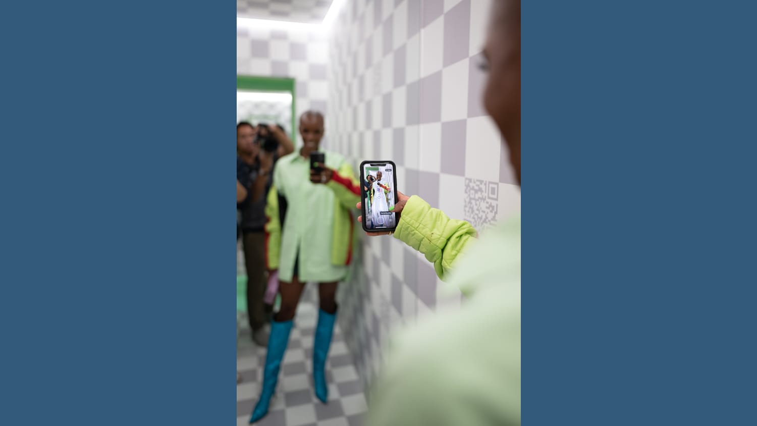 A black woman wearing a green shirtdress holds her phone up to a mirror. In her phone screen, she tries on a white digitally rendered dress.