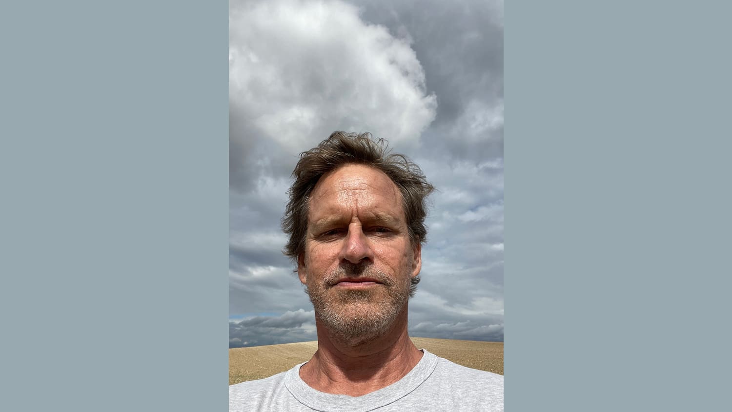 Photo of a man with blonde hair and a beard, looking into the camera. Behind him: a hilly field and storm clouds.