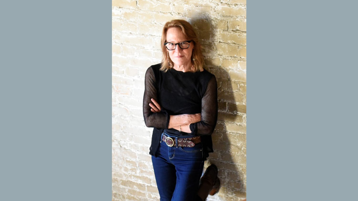 A photo of a woman with blonde hair leaning up against a beige brick wall. She stands with her arms crossed wearing glasses, a black mesh quarter-sleeve shirt and jeans.