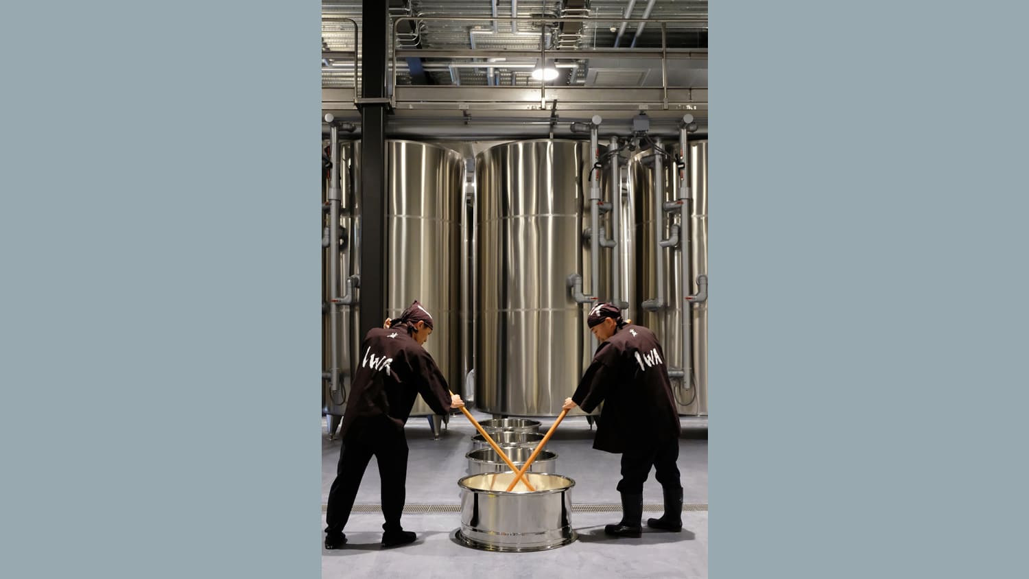 Two men wearing all black lean over a steel barrel partially underground, holding long wooden pegs and stirring within the barrel. Several large steel barrels stand in the background.