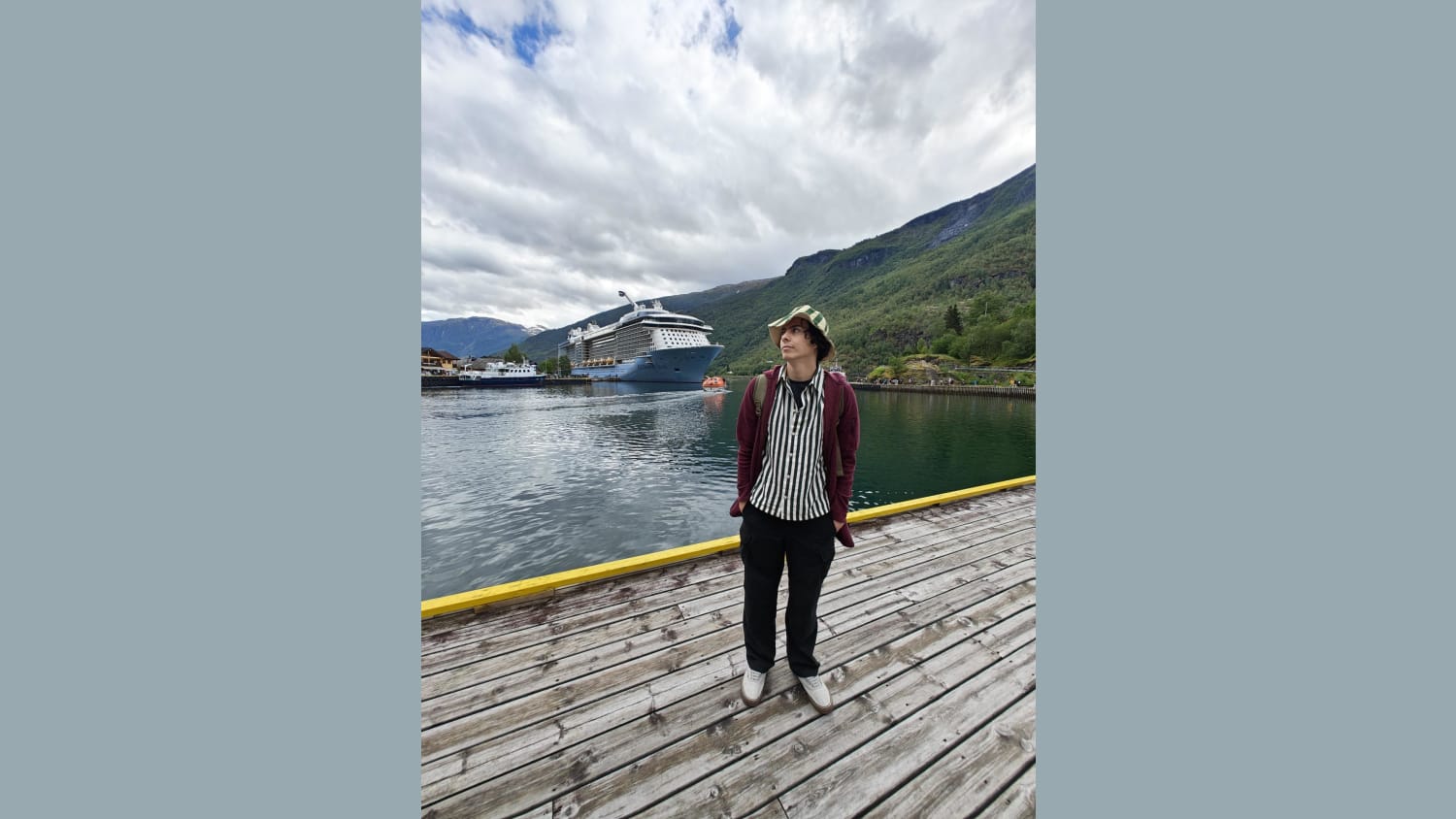 A man wearing a striped shirt, maroon jacket, and a striped bucket hat stands on a wooden pier with a yellow railing, looking up towards the sky. Behind him, a large blue and white cruise ship is docked in a body of water, flanked by towering green mountains under a cloudy sky.