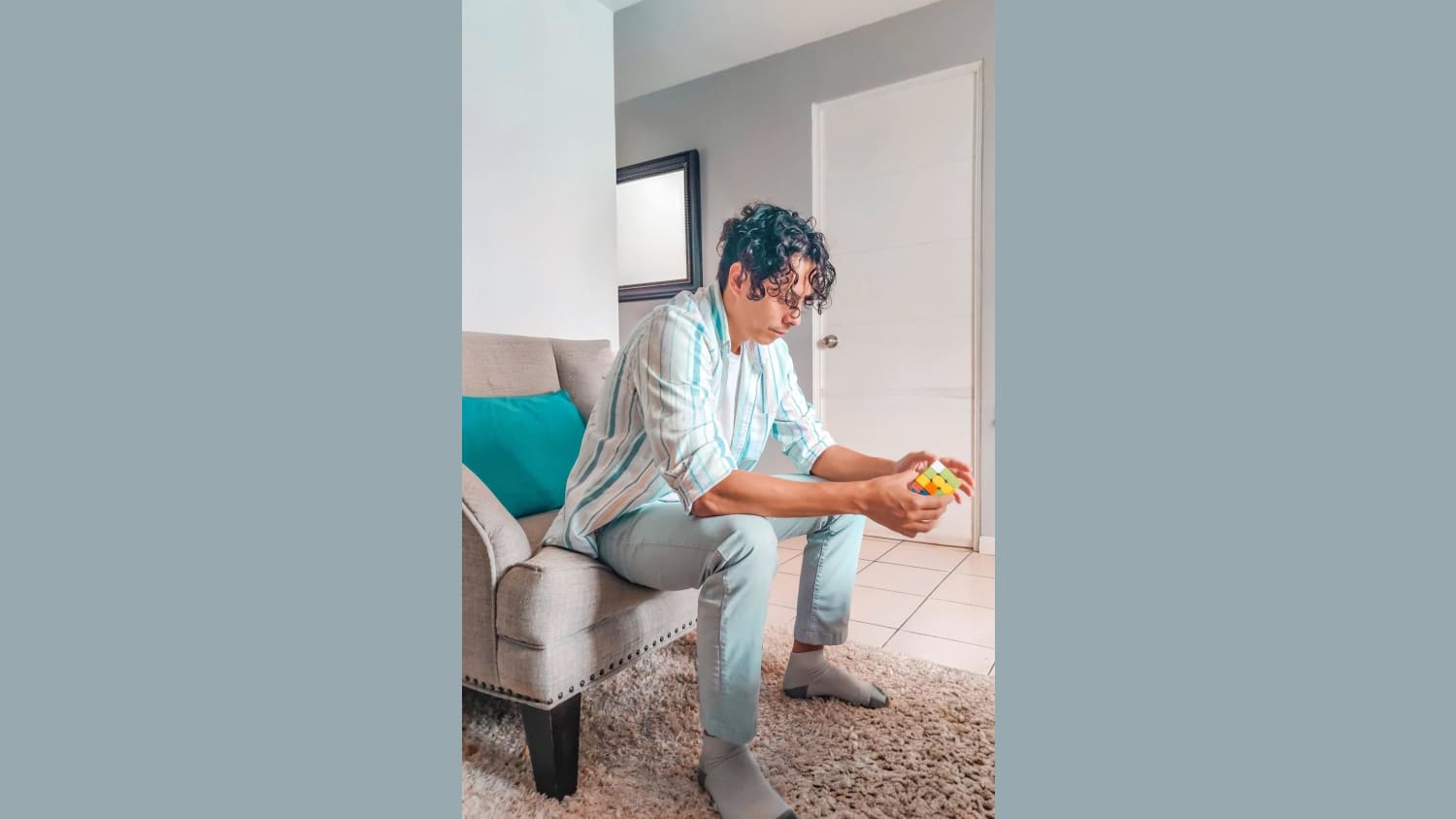 A young man with dark, curly hair sits on a beige armchair with a teal pillow, focused on a colorful Rubik's Cube in his hands. He is wearing a light-colored striped button-up shirt, light blue pants, and grey socks, seated on a textured beige rug in a room with white and gray walls and a white door.
