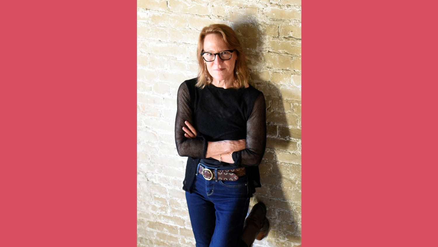 A photo of a woman with blonde hair leaning up against a beige brick wall. She stands with her arms crossed wearing glasses, a black mesh quarter-sleeve shirt and jeans.