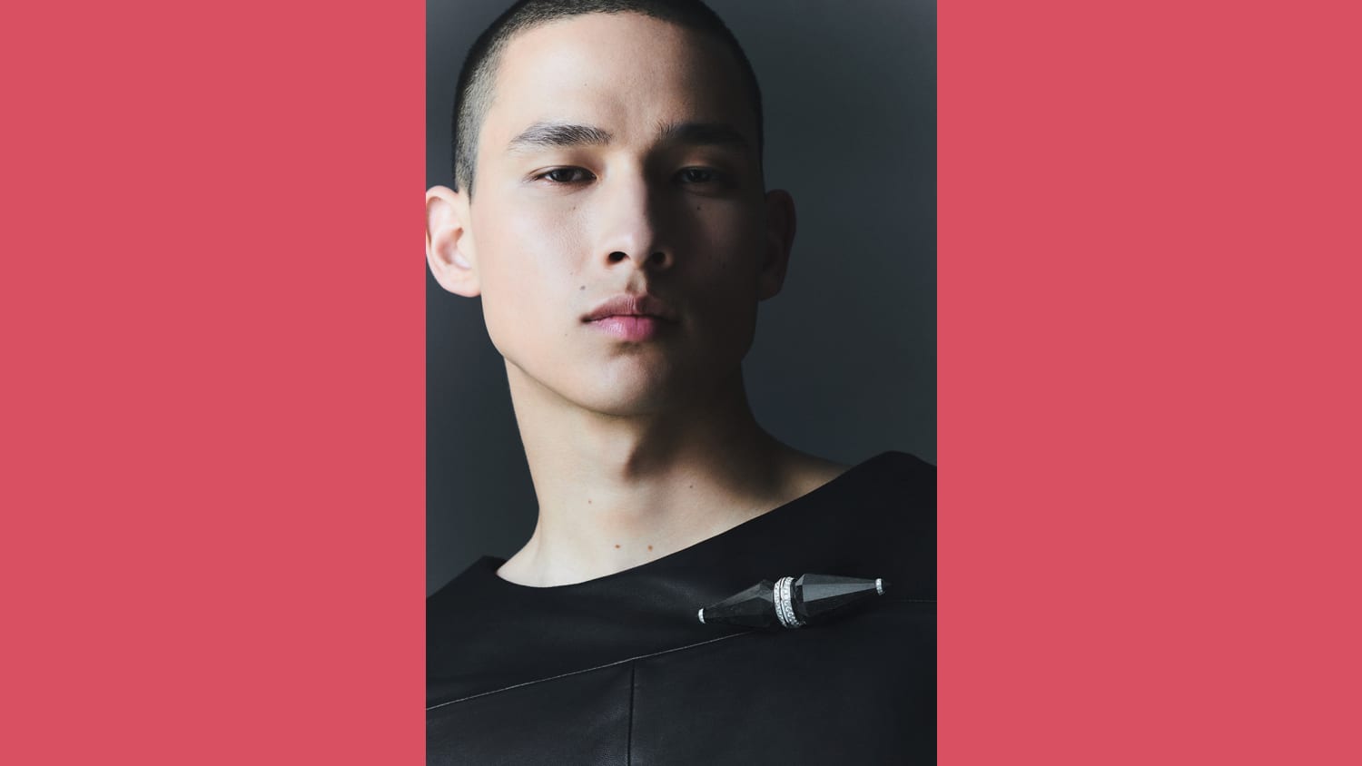 A male model with dark hair and light skin wears black in front of a black backdrop, wearing a black piece of jewelry pointed on both ends, with diamonds circling its center.