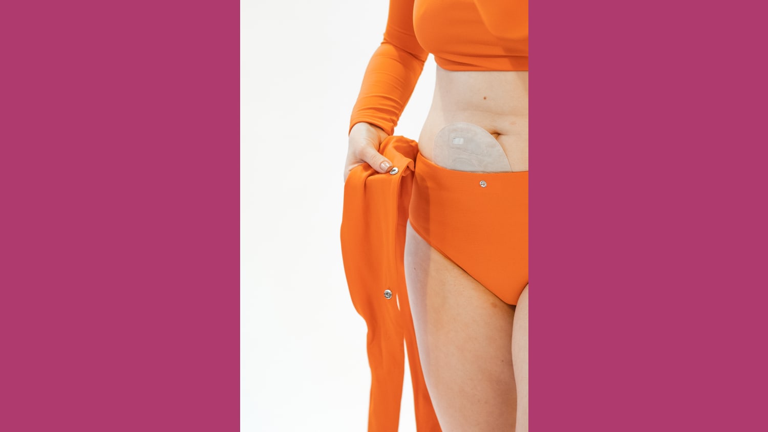 Close up on torso of a White woman with an ostomy bag who is putting on an adaptable bright orange two-piece swimsuit.