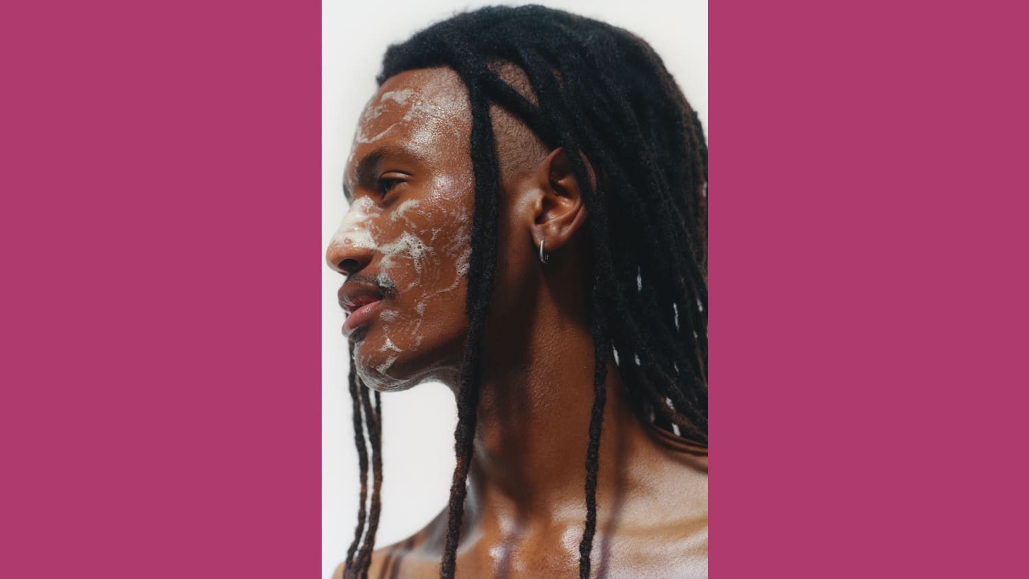 A black man with braided hair looks to the left of the screen, the image shows his chest-upwards. He shows one earring, has a thin mustache, and there is soap on his face.