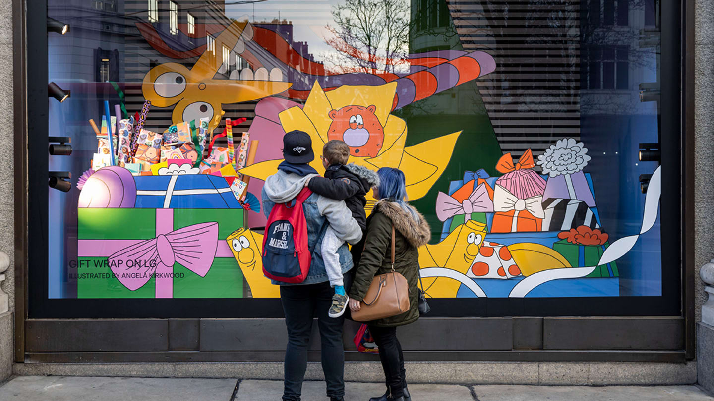 A man wearing a navy baseball cap holds a child and stands close to someone with blue hair. Their backs are to the viewer, as they look at a colorful window display from the street.
