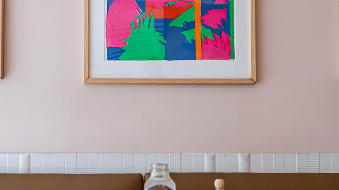 A table with one chair and one end on a booth, with a water pitcher, two glasses, tabasco sauce and salt and pepper shakers. A large, colorful painting hangs above the booth.