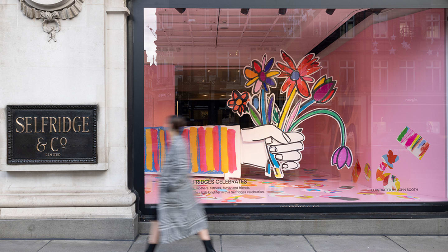 A woman blurred by movement walks by a pink window display of a giant cartoonish hand holding colorful giant cartoonish flowers. To the left, a sign reads Selfridge & Co.