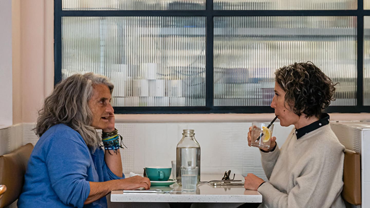 Two women sit at a small table in chairs in front of a blurred set of windows. The woman on the left has short gray hair and a blue shirt, and rests her head on her hand. On the right, a woman with short curly dark hair sips from a glass.