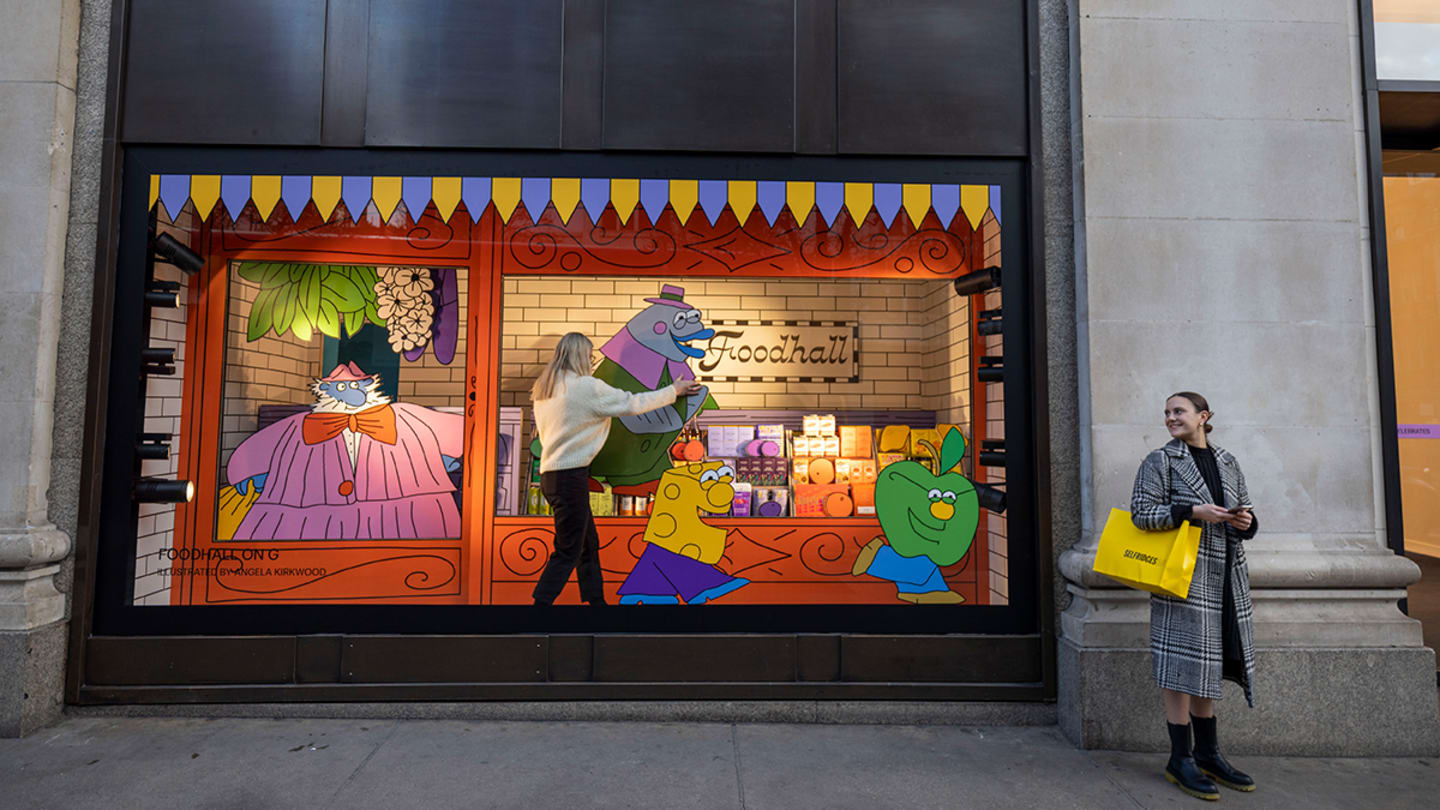 A woman with blonde hair is blurred in movement as she stands inside a colorful window display adjusting its contents: a cartoonish character with blue skin and an orange suit serves other cartoon characters. Another woman stands just outside the window.
