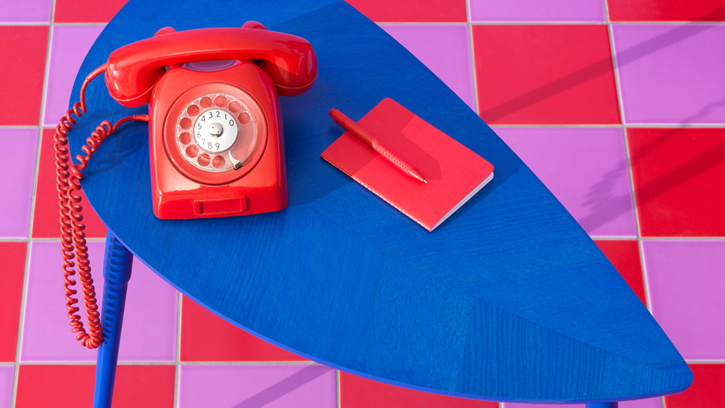 A blue teardrop shaped table holds a red telephone, notepad and pen above a light and dark pink checkered floor.