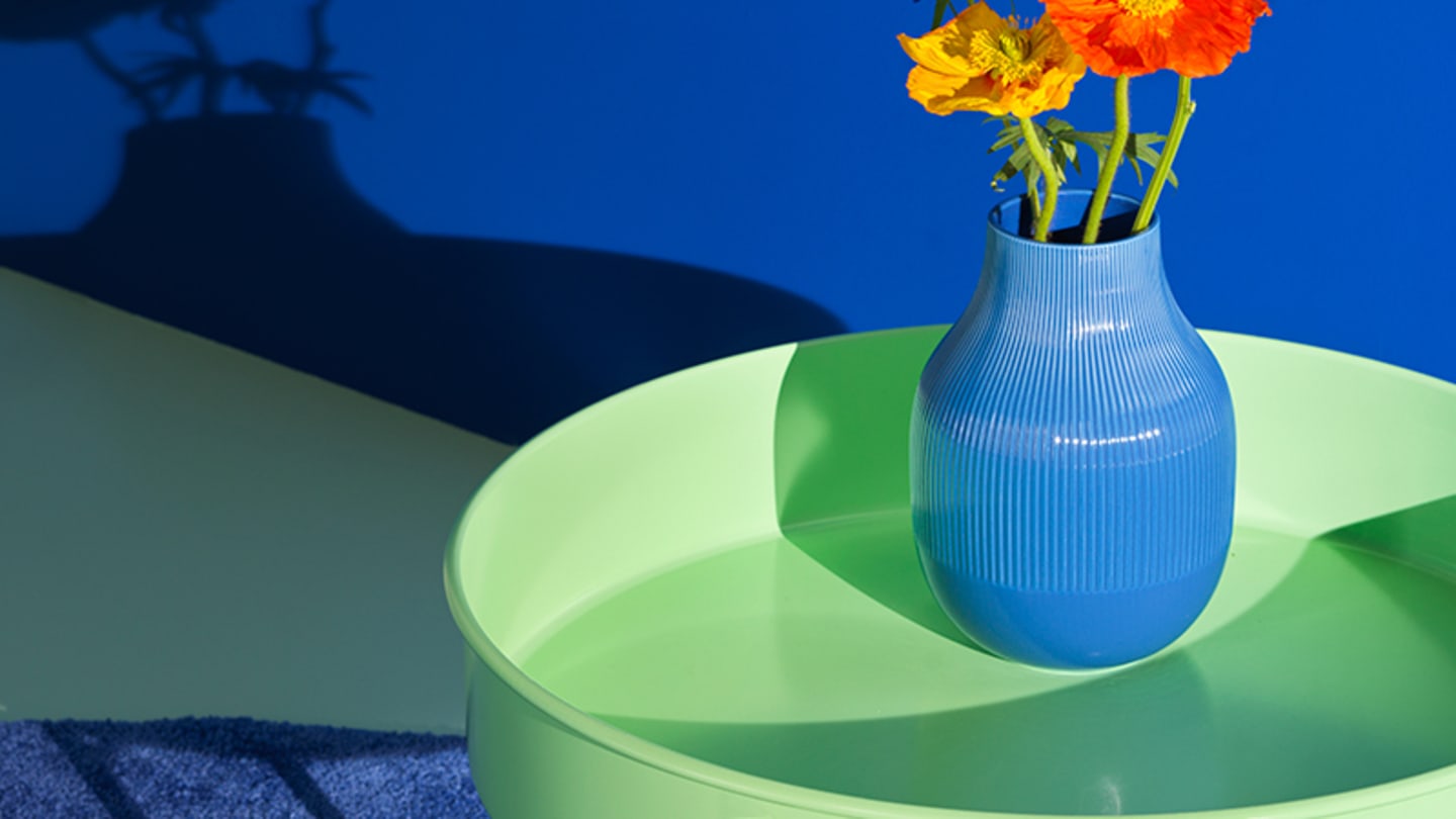 A circular lime green table with a blue transparent vase and orange flowers in a room with a dark blue wall.
