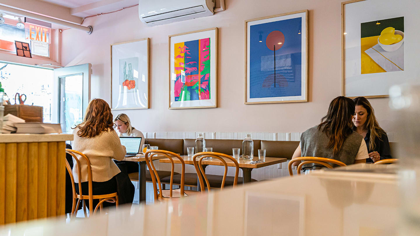 A set of 4 small tables, with two people sitting at the farthest table on the left and one person sitting at the farthest on the right, in front of four colorful paintings hung on the wall above a booth.