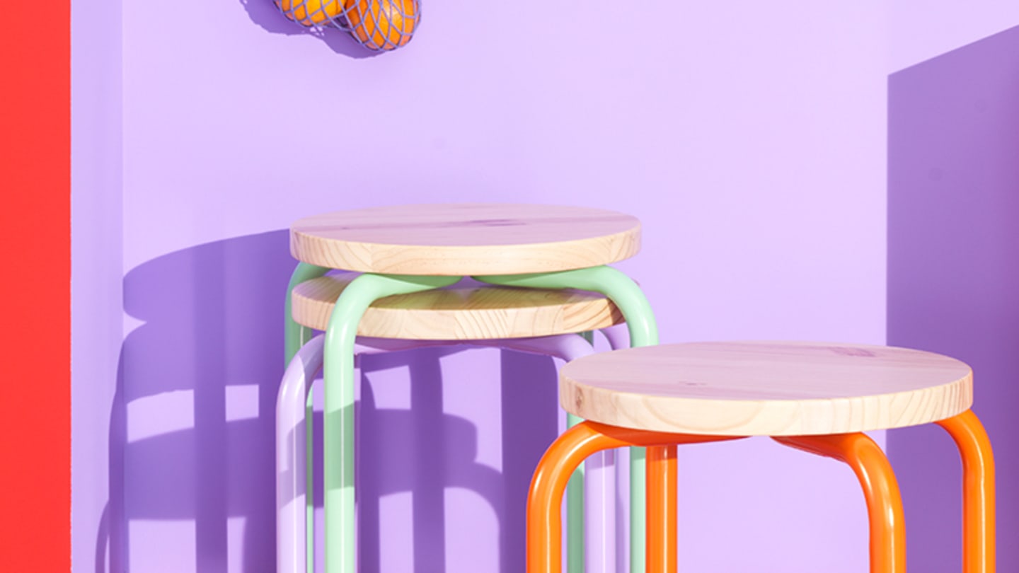 In a room with light link walls and dark pink furnishings, a purple mesh bag with two oranges and a pink hat hang above three stools, two stacked and one separate with orange legs.