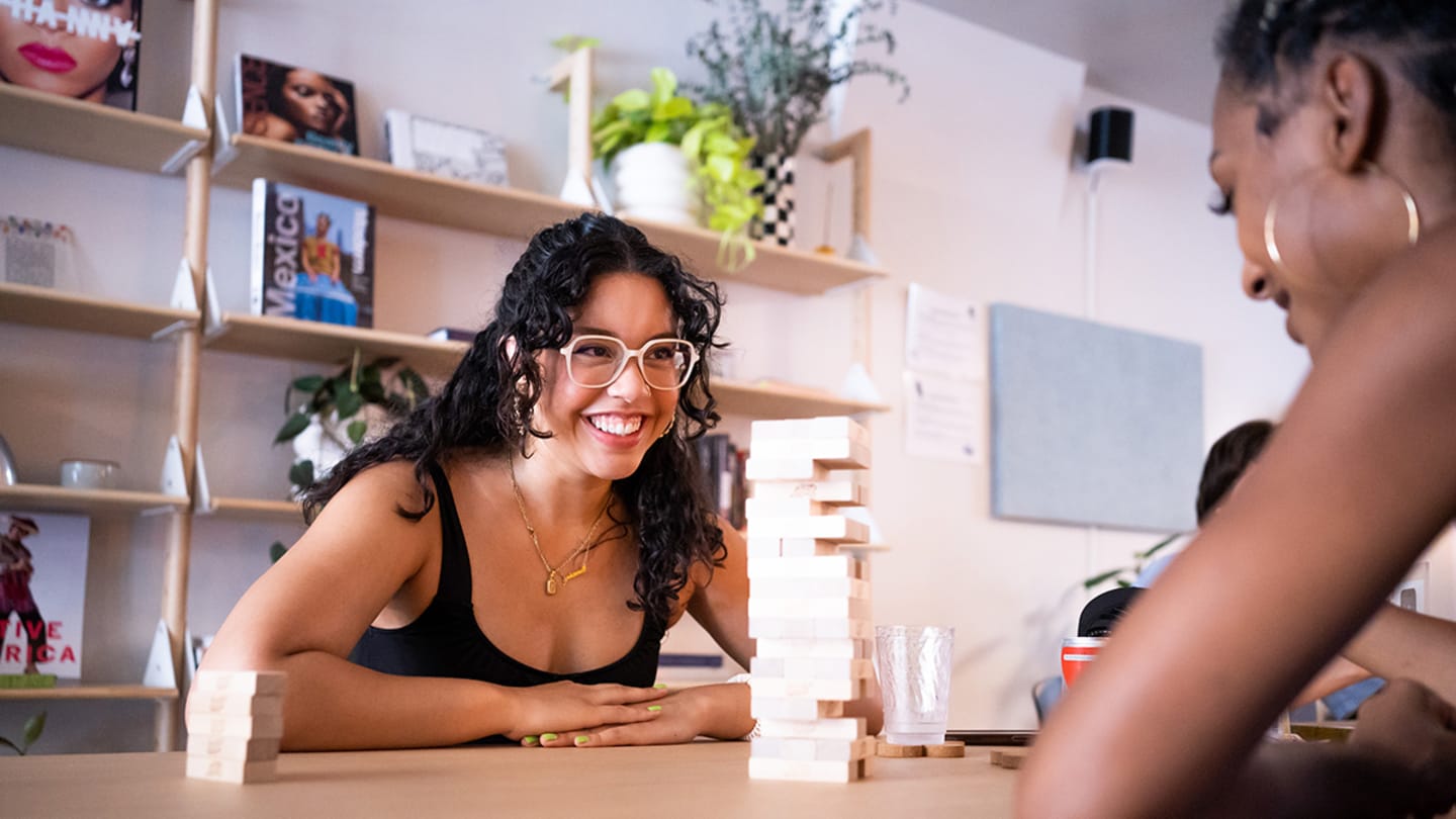 Two women with tan skin sit across from each other at a wooden table. The woman across the table, whose face is visible, has dark curly hair, glasses, and is smiling.