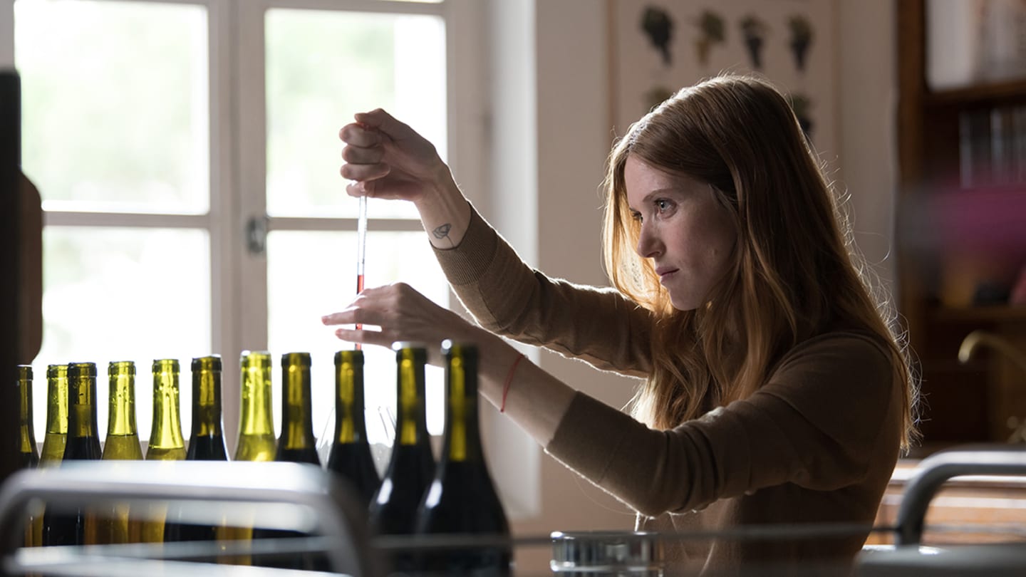 A woman with long light red hair and fair skin holds a dropper over a glass wine bottle.
