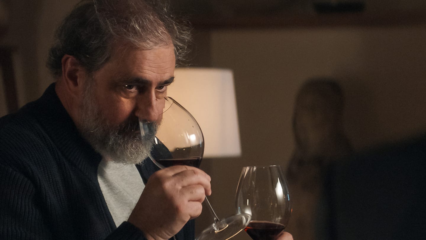 A man with dark graying hair looks to to the right of the camera, holding two glasses of wine. He sniffs one.