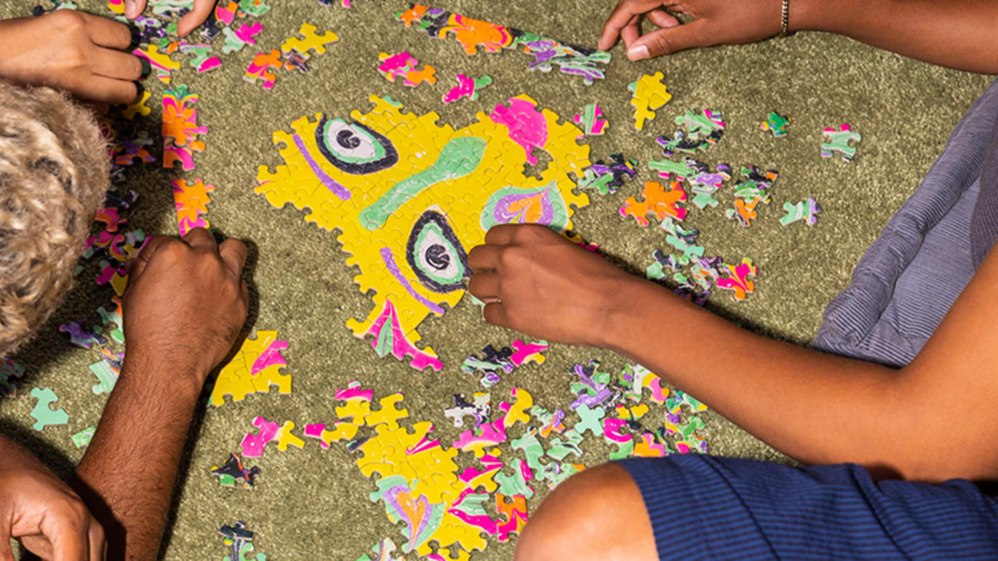 Two people with dark skin sit on a rug and work on a colorful, face-like puzzle.
