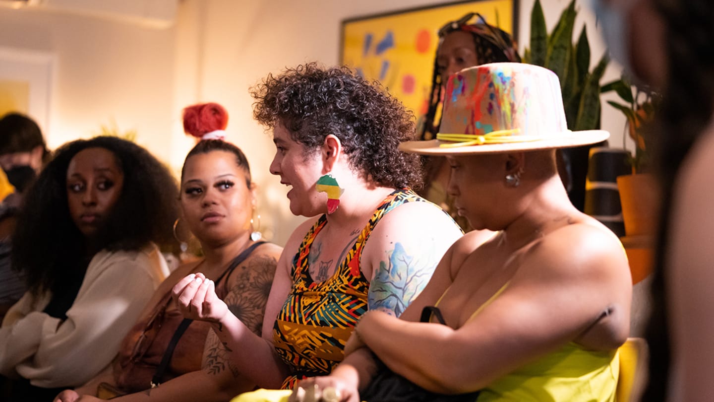 Five women sit on a couch, side by side. The second woman from the front is speaking. The woman closest to the viewer is wearing a colorful hat.