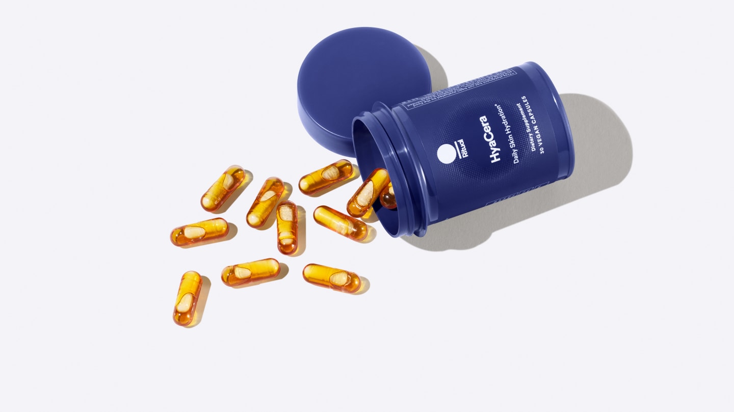 Dark blue cyclindrical pot lying on its side with yellow capsules filled with oil spilling across a white surface The label reads Hya Cera Daily Skin Hydration
