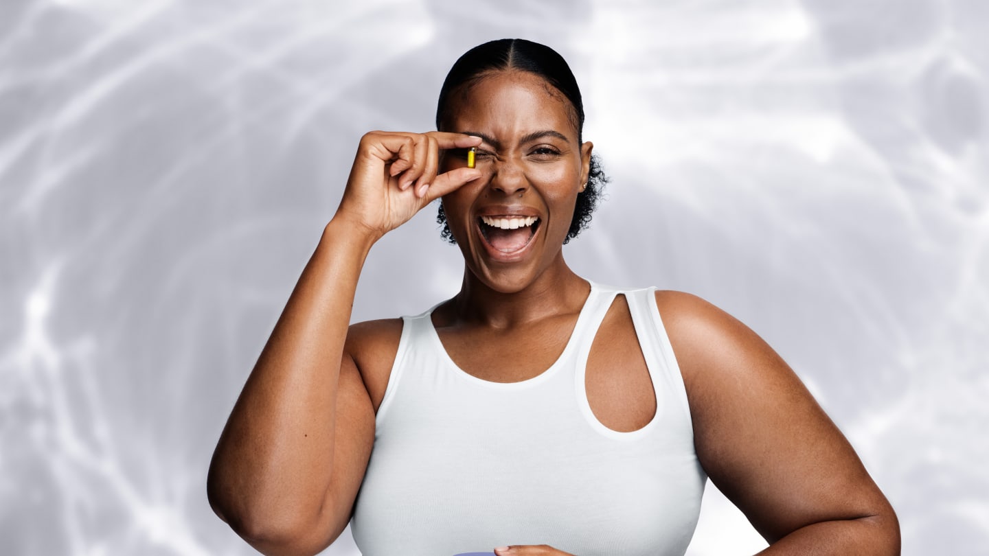 Black woman wearing a white formfitting vest with raindrop cutout at shoulder She is sat at a table with shiny surface and holding a yellow capsule in front of her right eye and smiling