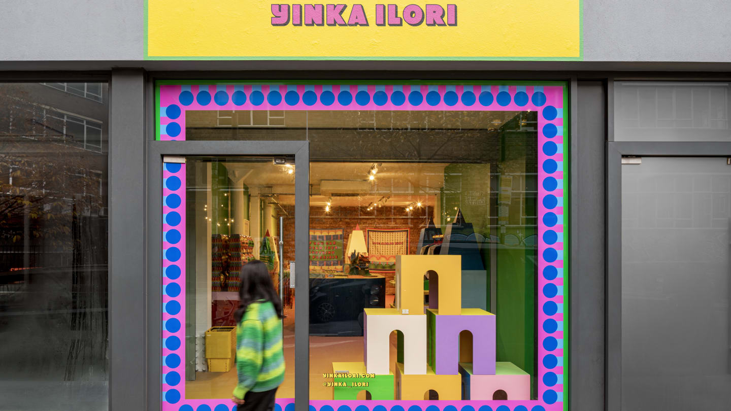 A woman in a green top walks past a store exterior A series of multicolored arch shapes are in the window which is bordered with colored trim Above the store is the sign saying Yinka Ilori