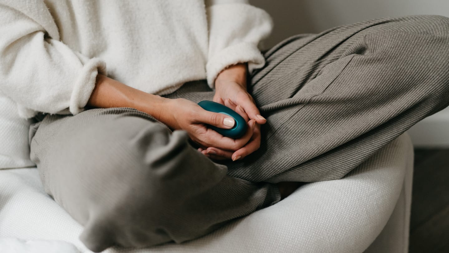 The lower half of a white woman in light trousers and a soft cream sweater holding a green pebble shaped device in her hand