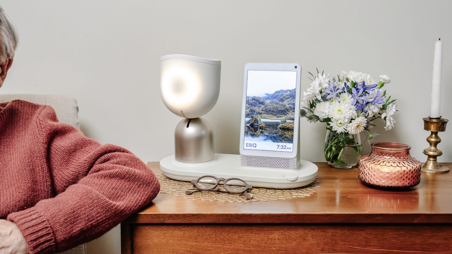 A white lamp shaped robot device with a small screen sits on top of a wooden dresser On the left we can see the sleeve of its edlerly owner