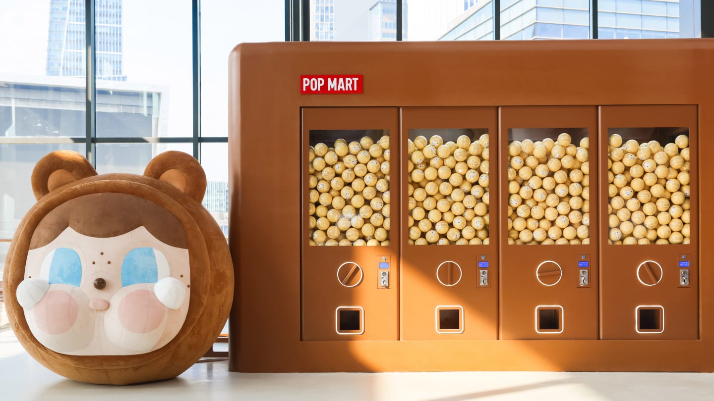 Giant brown plushy bear-face beside a POP MART display case filled with small boxed figures in front of windows.