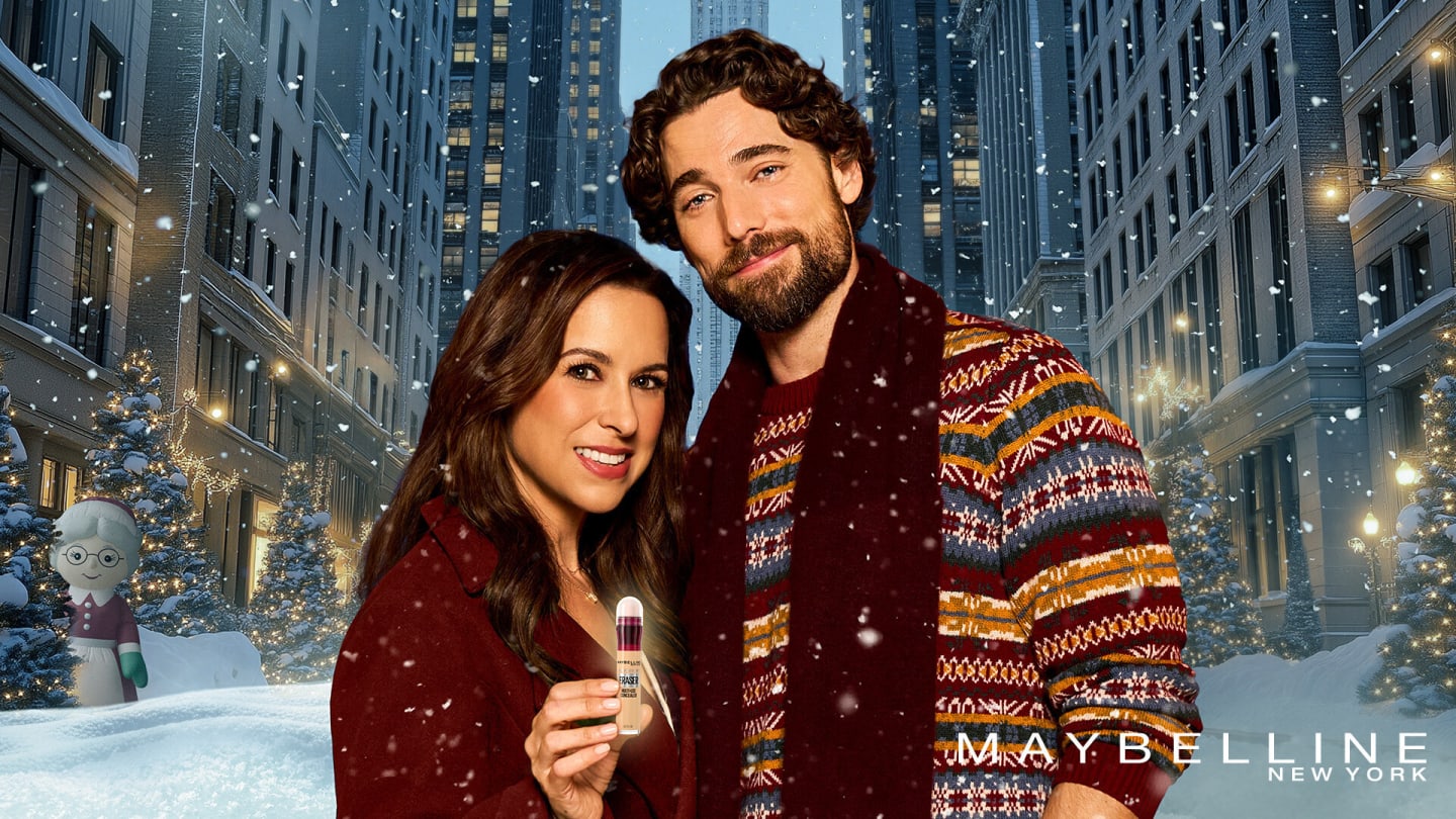 A dark-haired couple stand in the snow smiling, against a New York backdrop. The woman is holding a Maybelline foundation bottle.