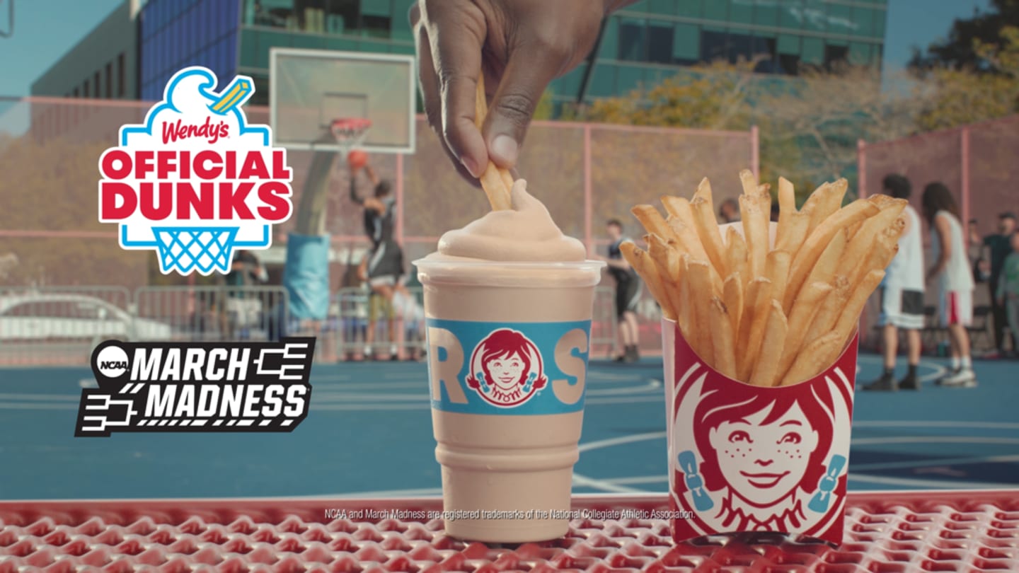 A person dips a fry into a Wendy’s Frosty beside a fries container. "Wendy's Official Dunks" and "NCAA March Madness" logos are visible. Basketball game in the background.
