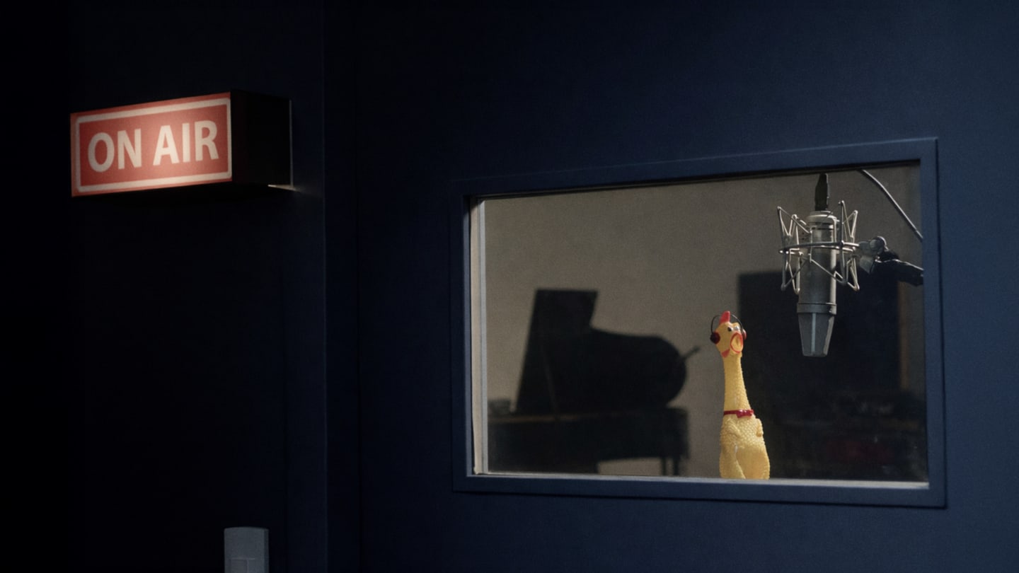 A rubber chicken in a sound booth speaking into a microphone with an on air sign visible.